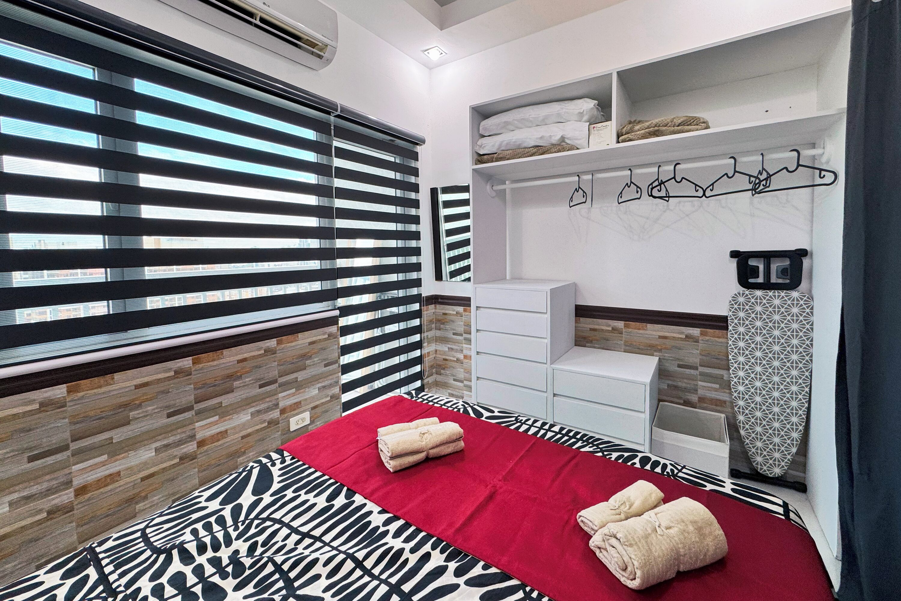 A modern hotel room with a patterned bedspread, a red blanket, a neatly made bed with towels, a white wardrobe, a hanging clothes rack, and a window with blinds.