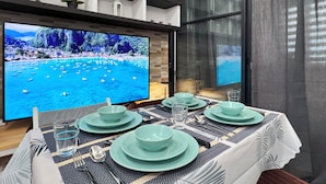 Un coin repas avec une table dressée pour quatre personnes, un grand téléviseur à écran plat diffusant une scène de plage et un aménagement intérieur moderne.