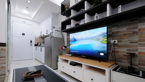 Un salon moderne avec un téléviseur à écran plat, un meuble de rangement en bois, des armoires blanches et un coin cuisine à l’arrière-plan.
