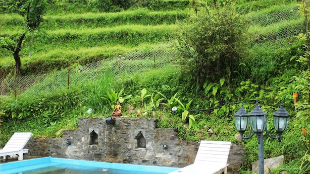 Una piscina con sillas de descanso blancas, un muro de piedra con estatuas y colinas verdes exuberantes al fondo.