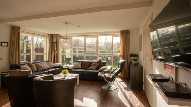 A spacious living room with a dark-colored sofa set, a modern chair, and a coffee table with a plant. Large windows provide natural light and a view of the outdoors. - Duinweg 12 comfortable luxury holiday villa near beach (Renesse)