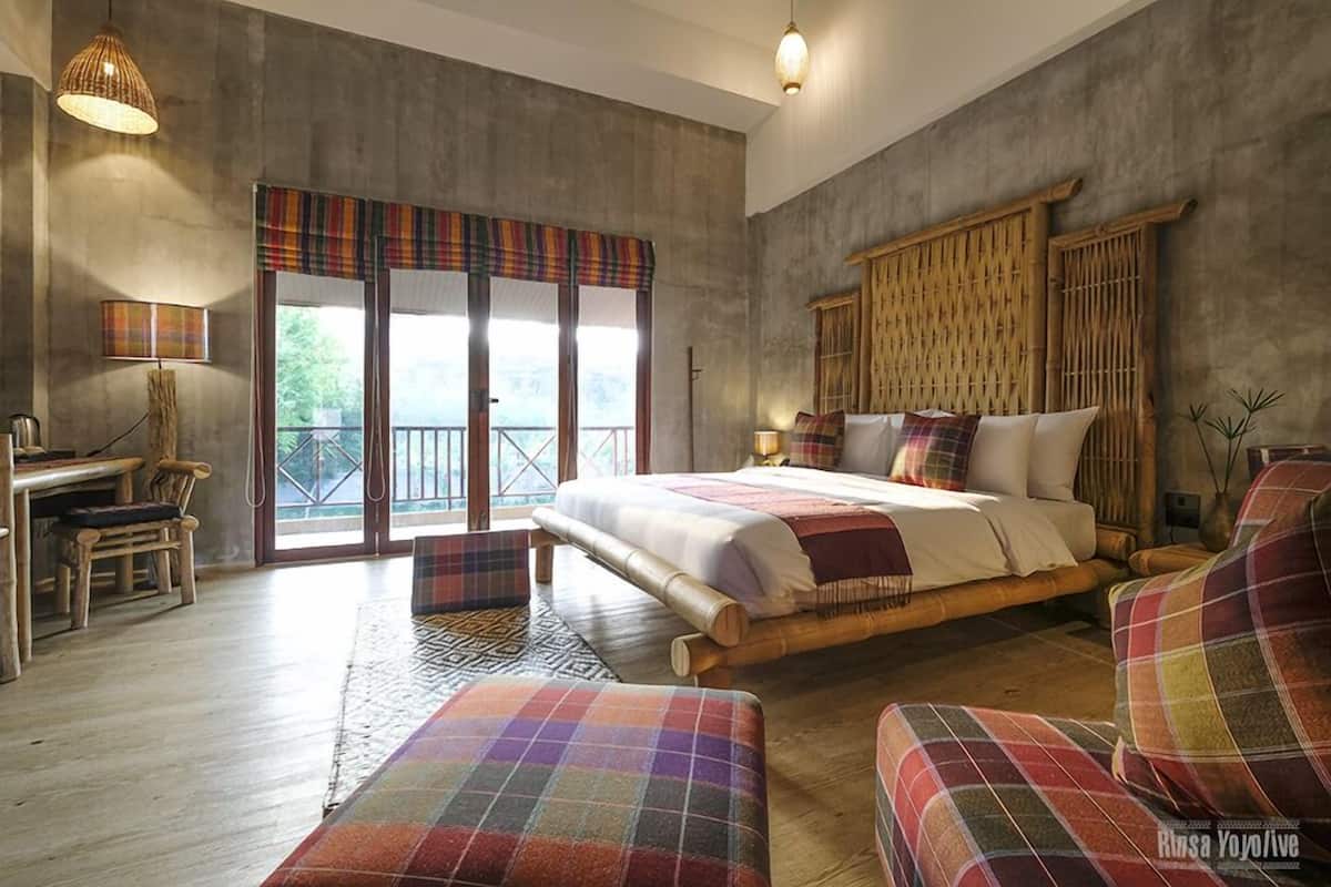 A bedroom with a large bed, a desk, and a chair. There is a window with a view of greenery outside.