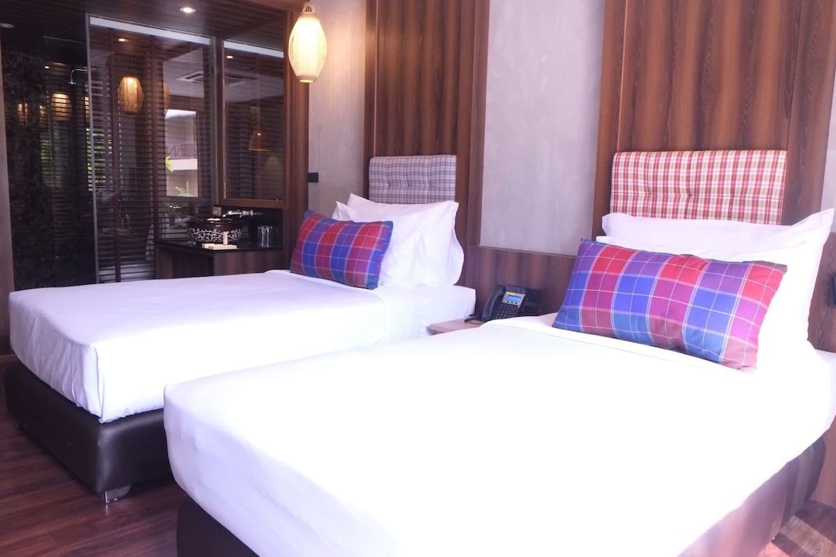 A hotel room with two beds, each with white linens and plaid pillows. There is a desk with a telephone and a small table with items on it. The room has wooden paneling and a large window with blinds.