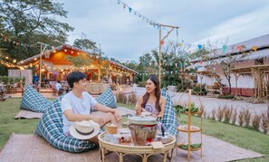 A couple sitting on a checkered cushion outdoors with a table in front of them, surrounded by thatched-roof structures and string lights. - Recall Isaan Isan Concept at Khaoyai (Pak Chong)