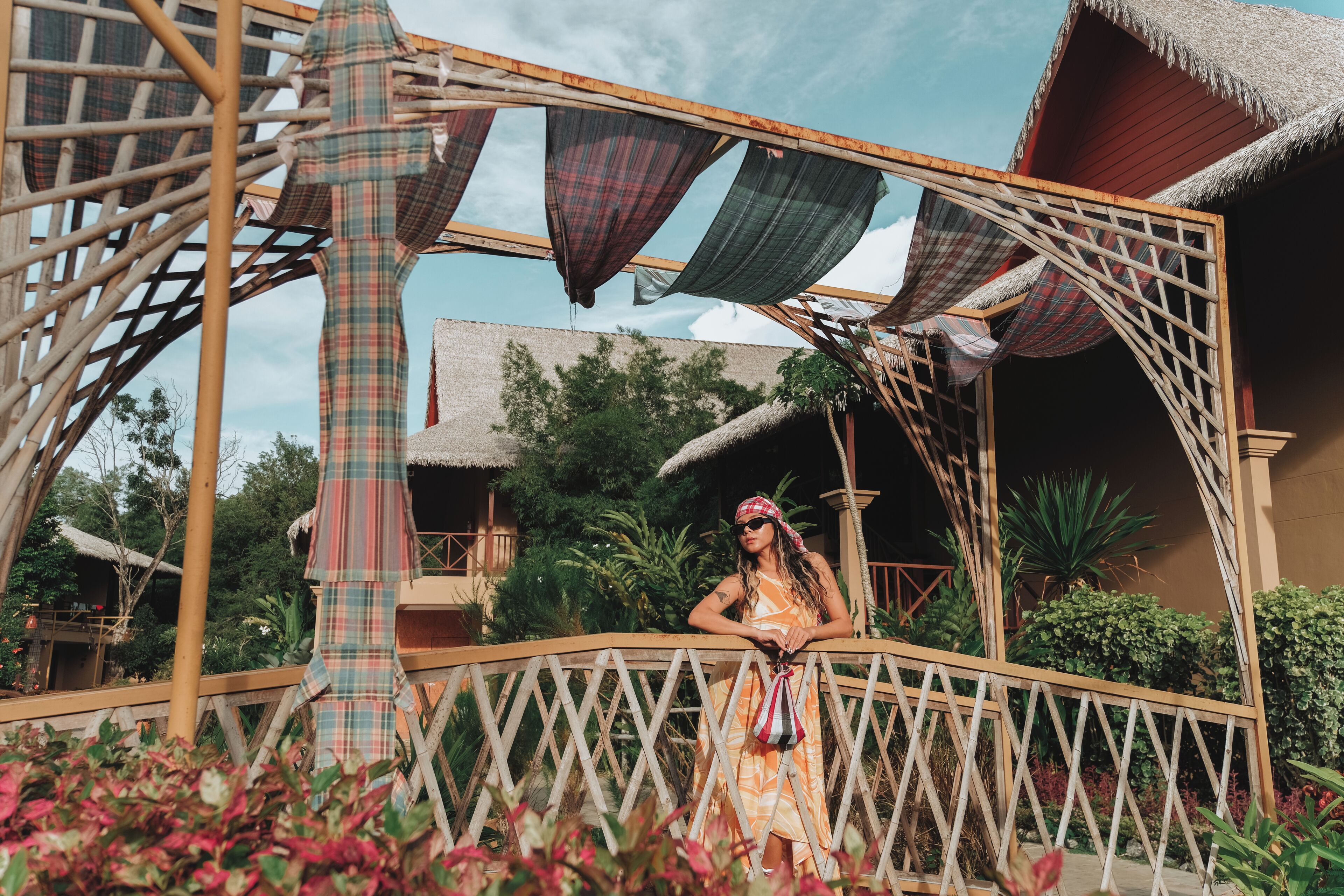 A person standing on a wooden bridge with a thatched roof structure, surrounded by lush greenery and a clear sky.