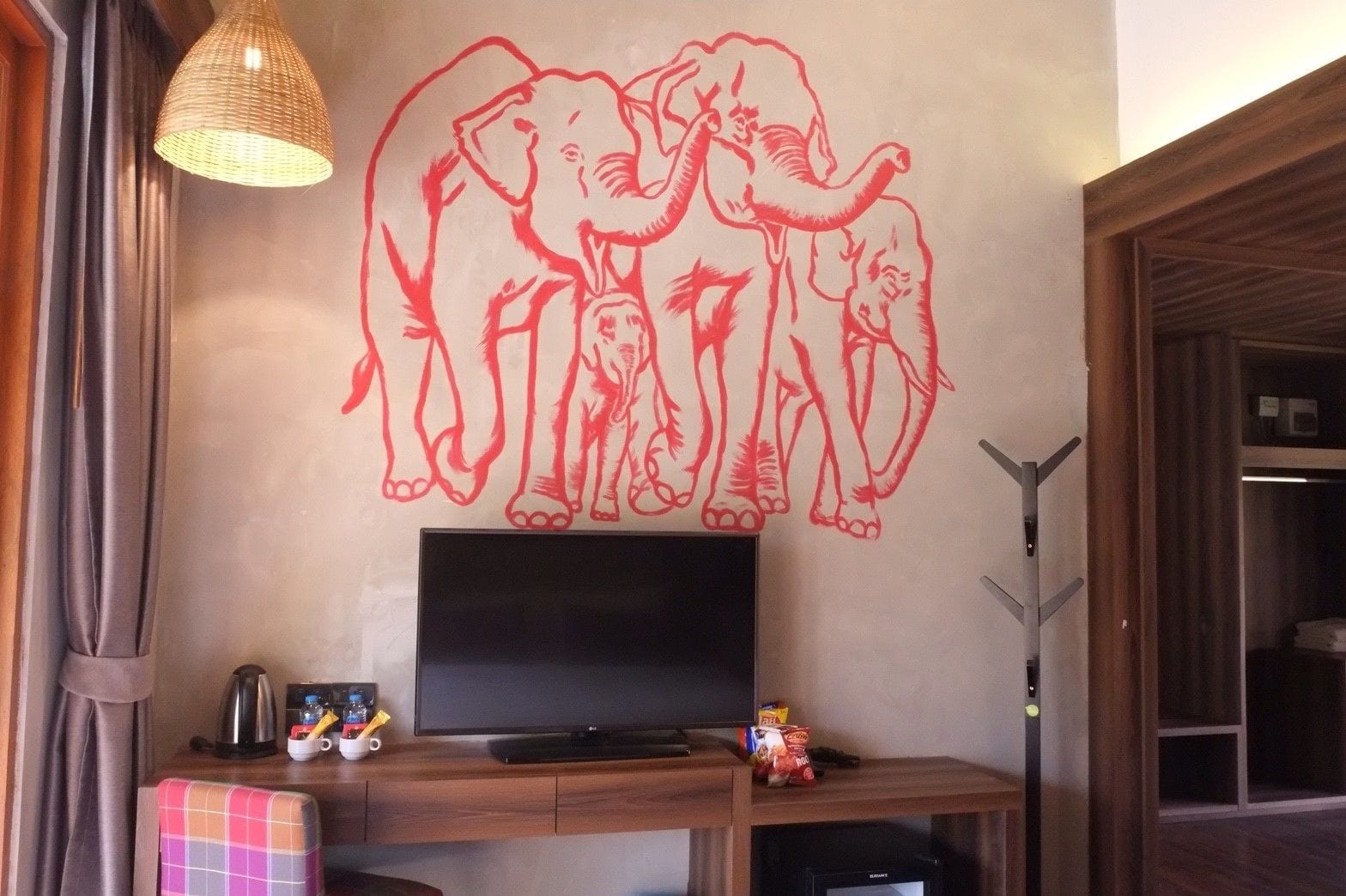 En væg med et stort vægmaleri af en rød elefant, et fladskærms-tv, en kaffemaskine og en lille hylde med snacks.