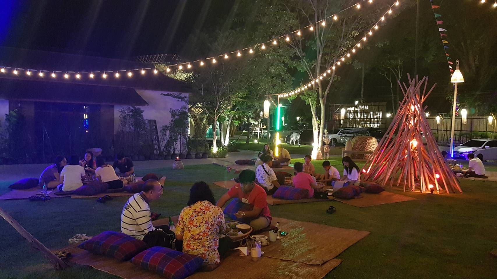 An outdoor gathering with people sitting on mats under string lights, a fire pit, and a building in the background.