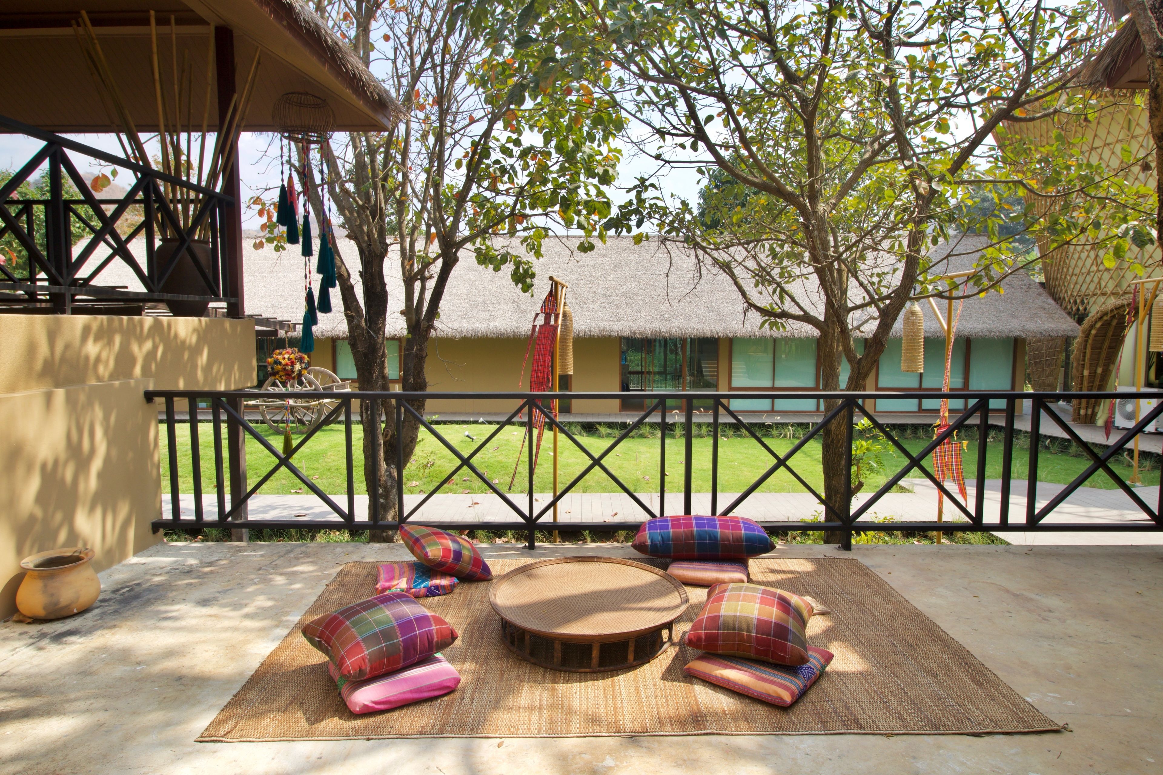 An outdoor seating area with woven mats and colorful cushions, a thatched-roof building in the background, and a black metal fence.