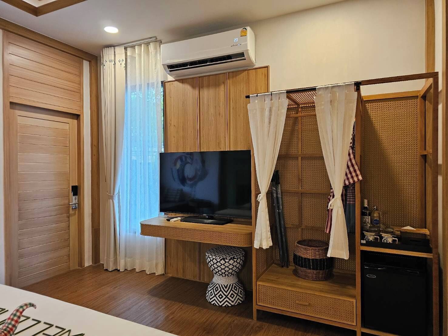 A room with a wooden wardrobe, a television on a wooden stand, a small black and white patterned stool, a wicker basket, and a black refrigerator.