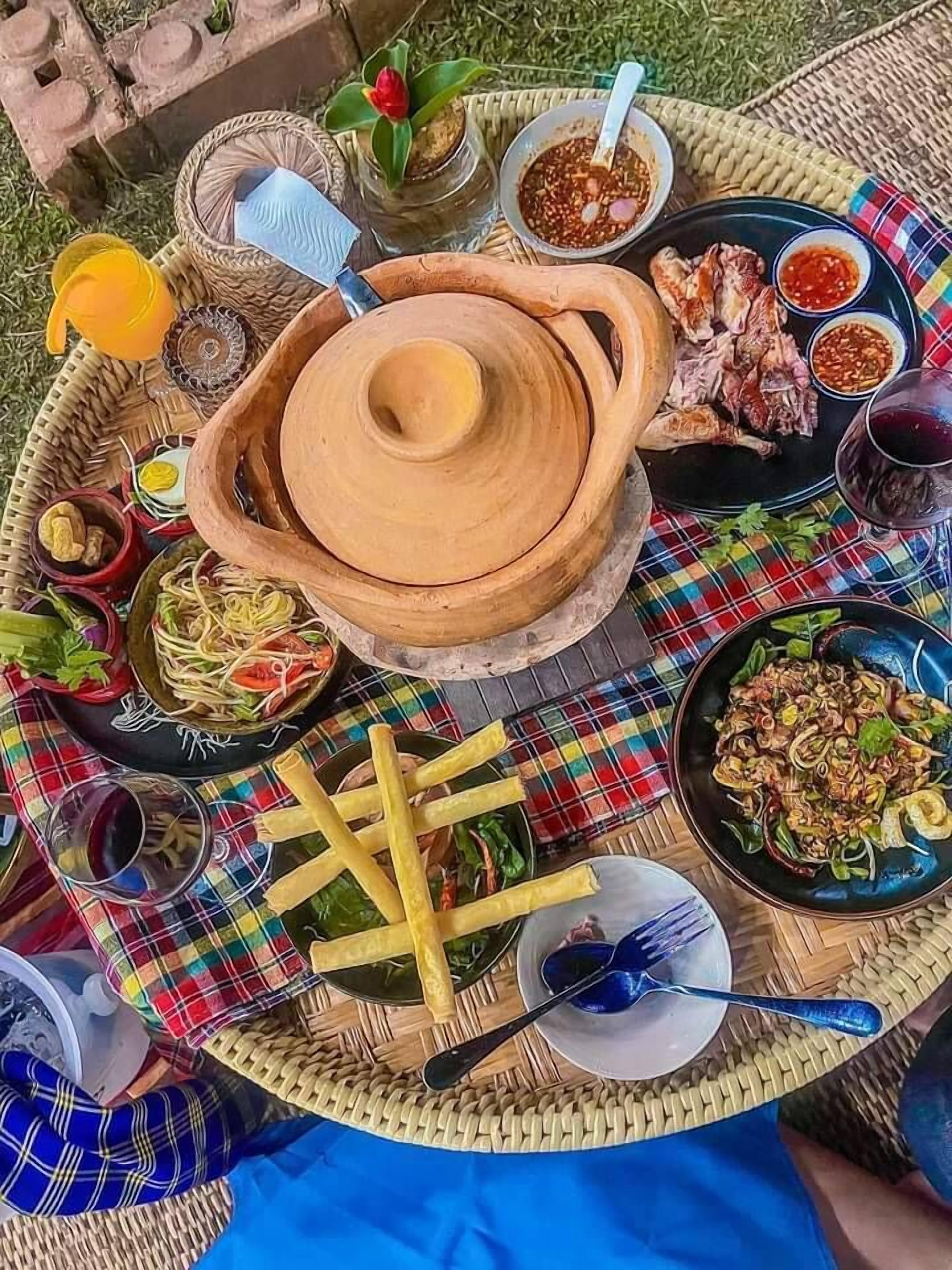 A variety of dishes and condiments on a woven tray, including noodles, fried items, and a large clay pot.