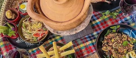 Una variedad de platos y condimentos sobre una bandeja tejida, entre ellos fideos, comida frita y una gran olla de barro.