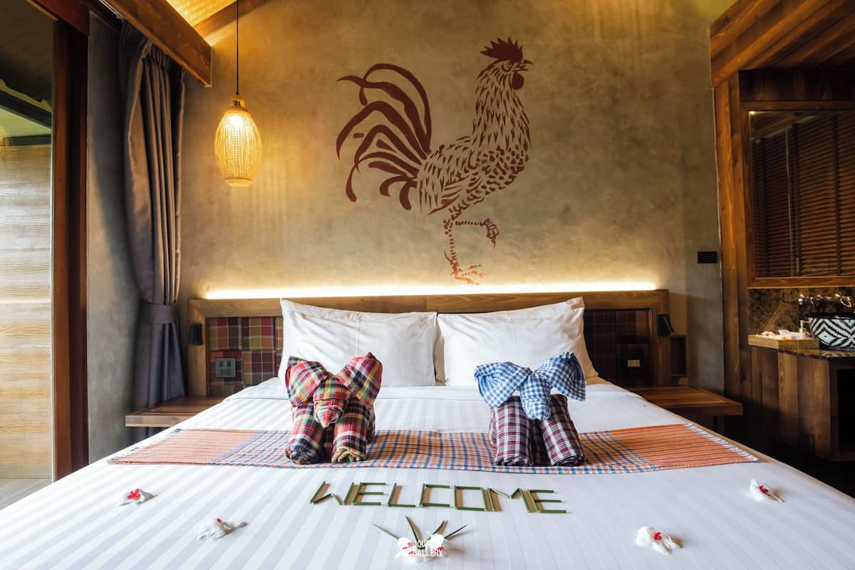 A hotel room with a large rooster mural on the wall, a neatly made bed with plaid bedding, and a welcome mat.