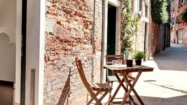 A narrow street with a brick wall on one side and a wooden table and chair set on the other. - Guggenheim Collection Venice Apartment (Venice)