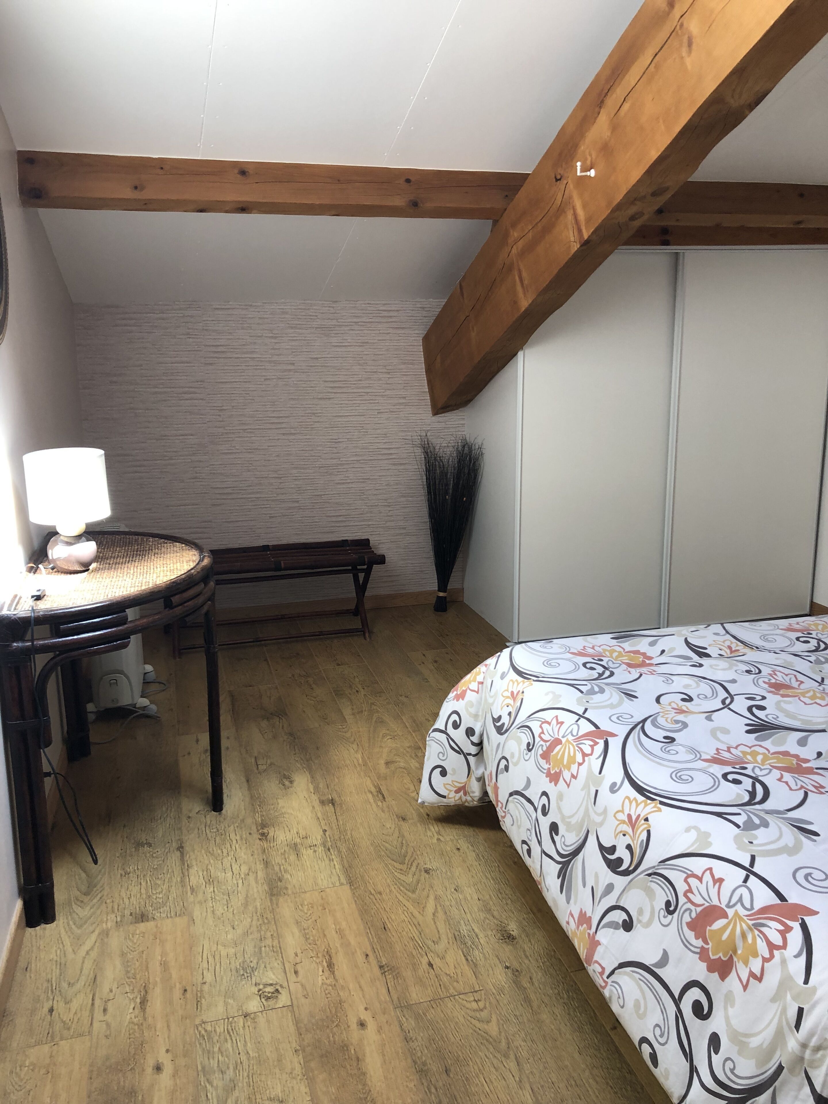 A bedroom with a wooden ceiling, a bed with a floral bedspread, a small table with a lamp, and a bench.