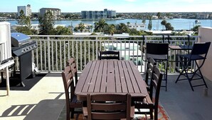 A balcony with a wooden table and chairs, a grill, and a view of a body of water and buildings in the distance. - 4 BR Clearwater Beach Townhouse • Hot Tub • Rooftop Lounge • Beach/Gulf Views (Clearwater)