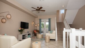 A spacious living area with a white sofa, a television, wicker side tables, and a staircase with a white railing. - 4 BR Clearwater Beach Townhouse • Hot Tub • Rooftop Lounge • Beach/Gulf Views (Clearwater)