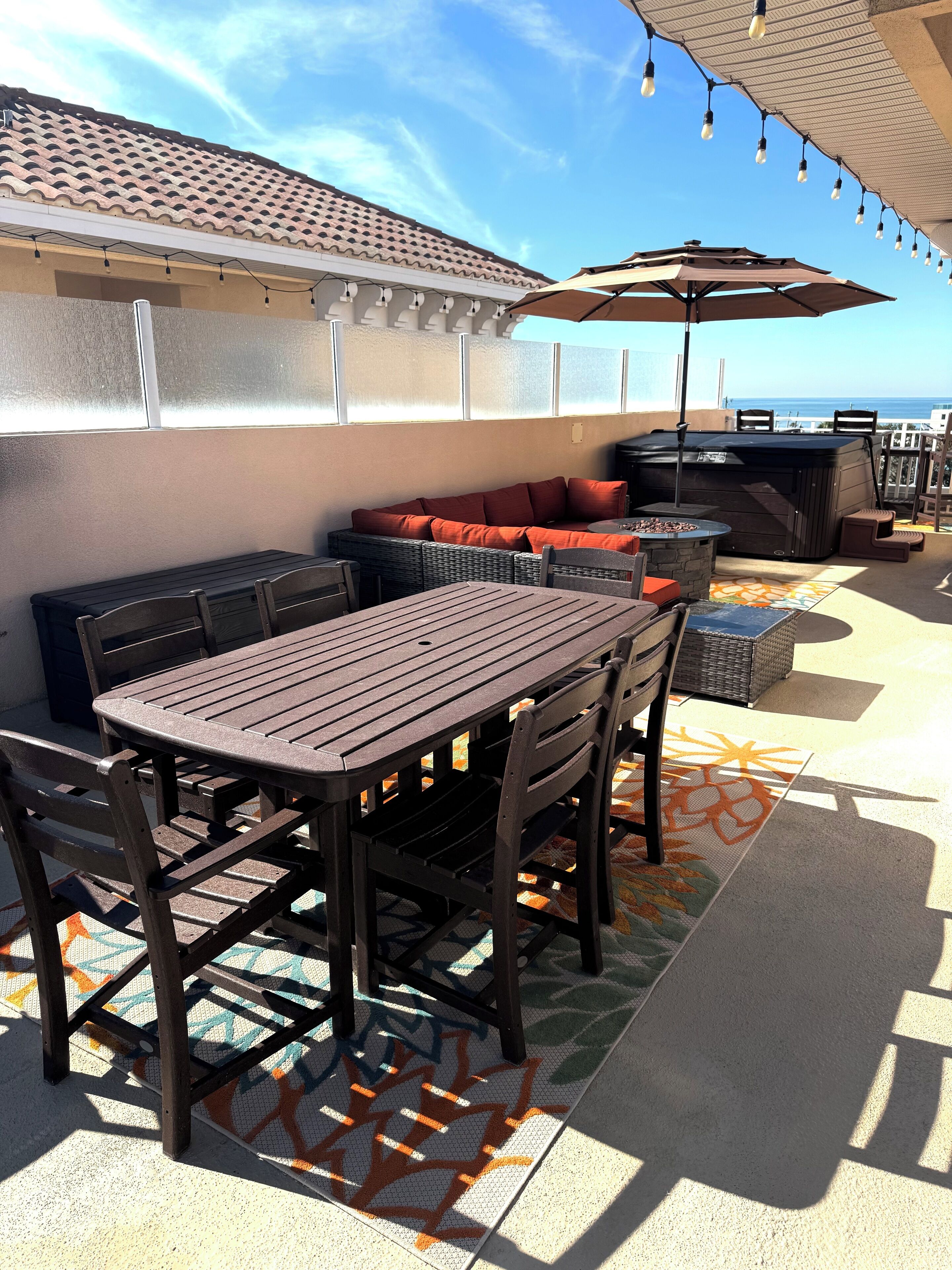 Une terrasse extérieure avec une table et des chaises, un coin salon confortable avec des coussins, un jacuzzi et des guirlandes lumineuses.