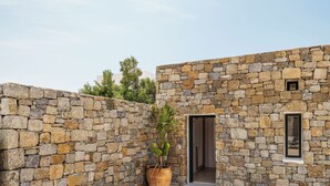 A stone-walled building with a large potted plant, a wooden bench, and a small window. - Mykonos Drops (Mykonos)