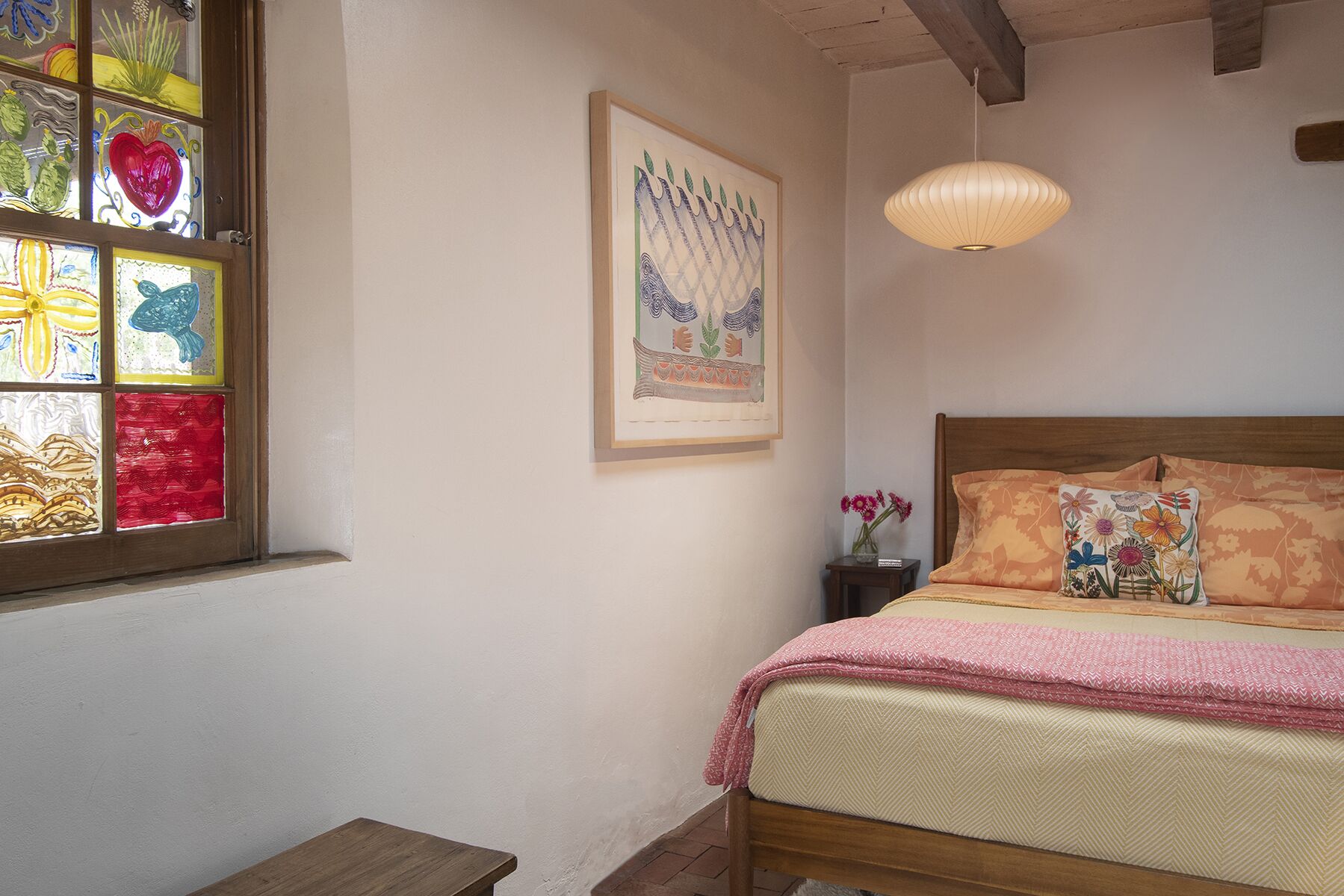 A bedroom with a bed, a nightstand, a hanging light, and a window with stained glass.