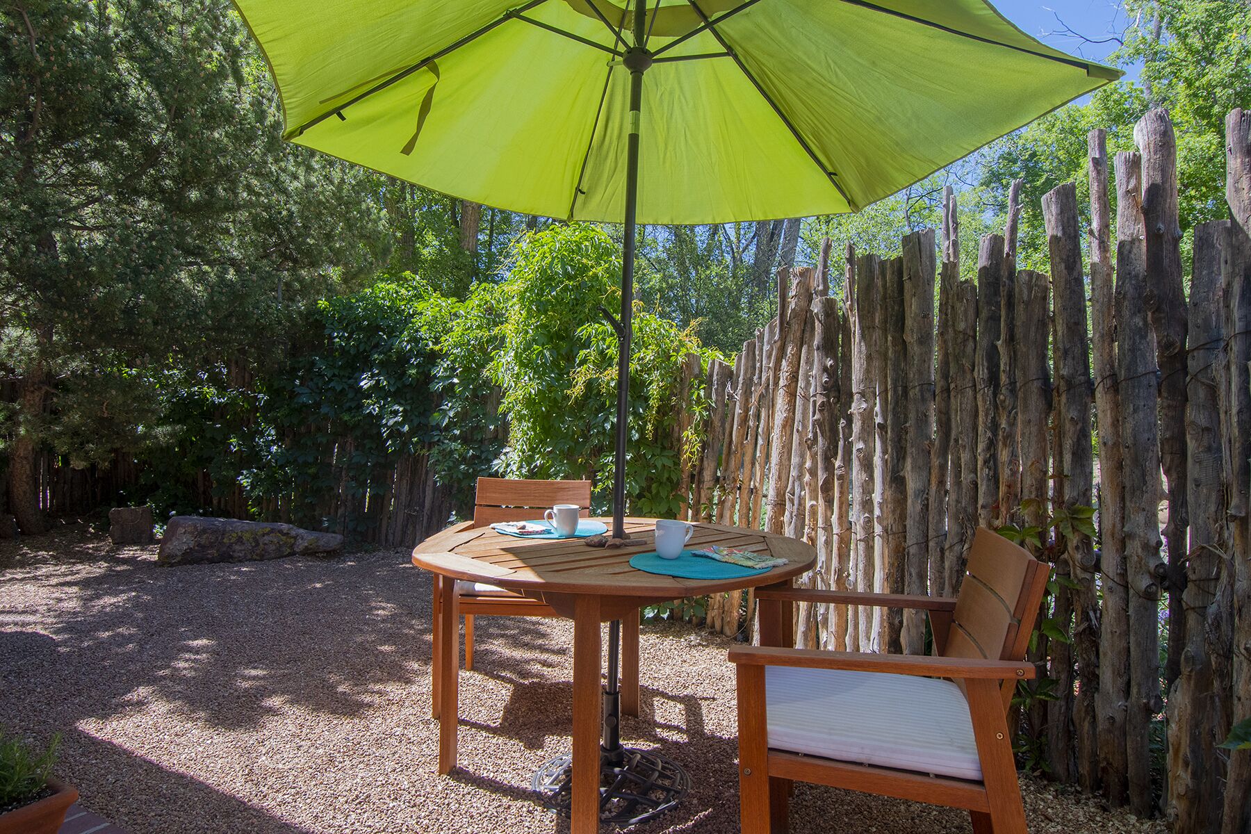 A wooden table and chairs under a green umbrella in a garden with a wooden fence.