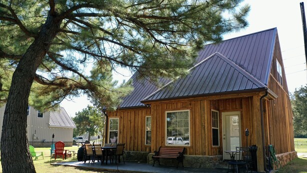 A wooden cabin with a metal roof, a porch with outdoor seating, and a large tree in the foreground. - Your Home Base for the Little Red River, Trout Fishing & Greers Ferry Lake (Heber Springs)