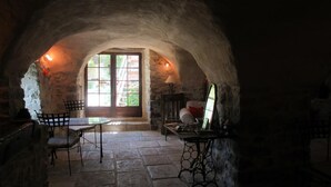 Ein Zimmer mit Steinwänden, ausgestattet mit einem Tisch, Stühlen, einem Kamin und einem Fenster mit Blick auf Grünflächen.