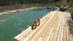 Eine Holzterrasse führt zu einer natürlichen Thermalquelle, umgeben von einem Holzzaun und üppiger Vegetation.