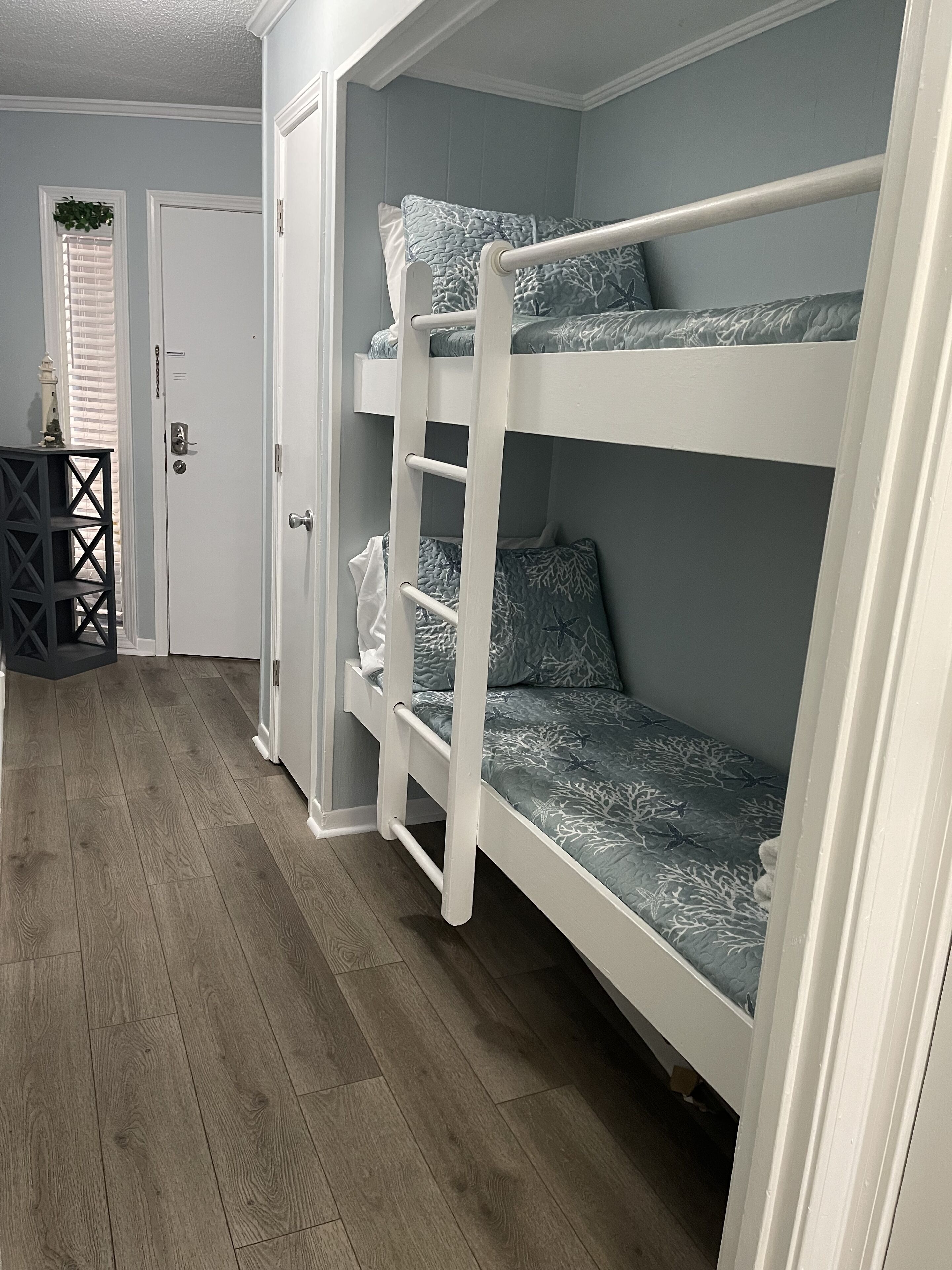 A bunk bed room with a wooden floor, a door with a keyhole, and a small table with a plant.
