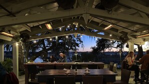 Un espace de restauration en plein air avec des tables et des chaises en bois, des guirlandes lumineuses et une vue sur les arbres et le coucher du soleil.