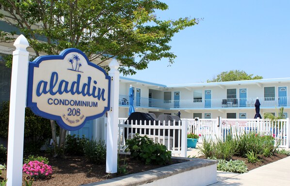 A sign for Aladdin Condominium 208 with a white picket fence and a pool area in the background.