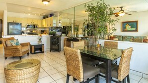 A modern living room with a glass dining table, wicker furniture, and a large mirror reflecting the kitchen area.