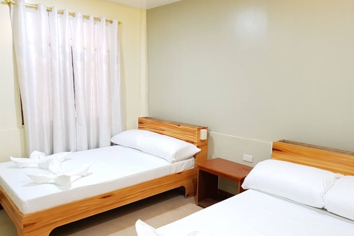 Ein Hotelzimmer mit zwei Holzbettgestellen, weißen Bettwäsche, einem Nachttisch aus Holz und einem Fenster mit weißen Vorhängen.