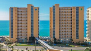 Un hôtel de grande hauteur doté de nombreux balcons, offrant une vue sur la plage et relié à un pont piétonnier.