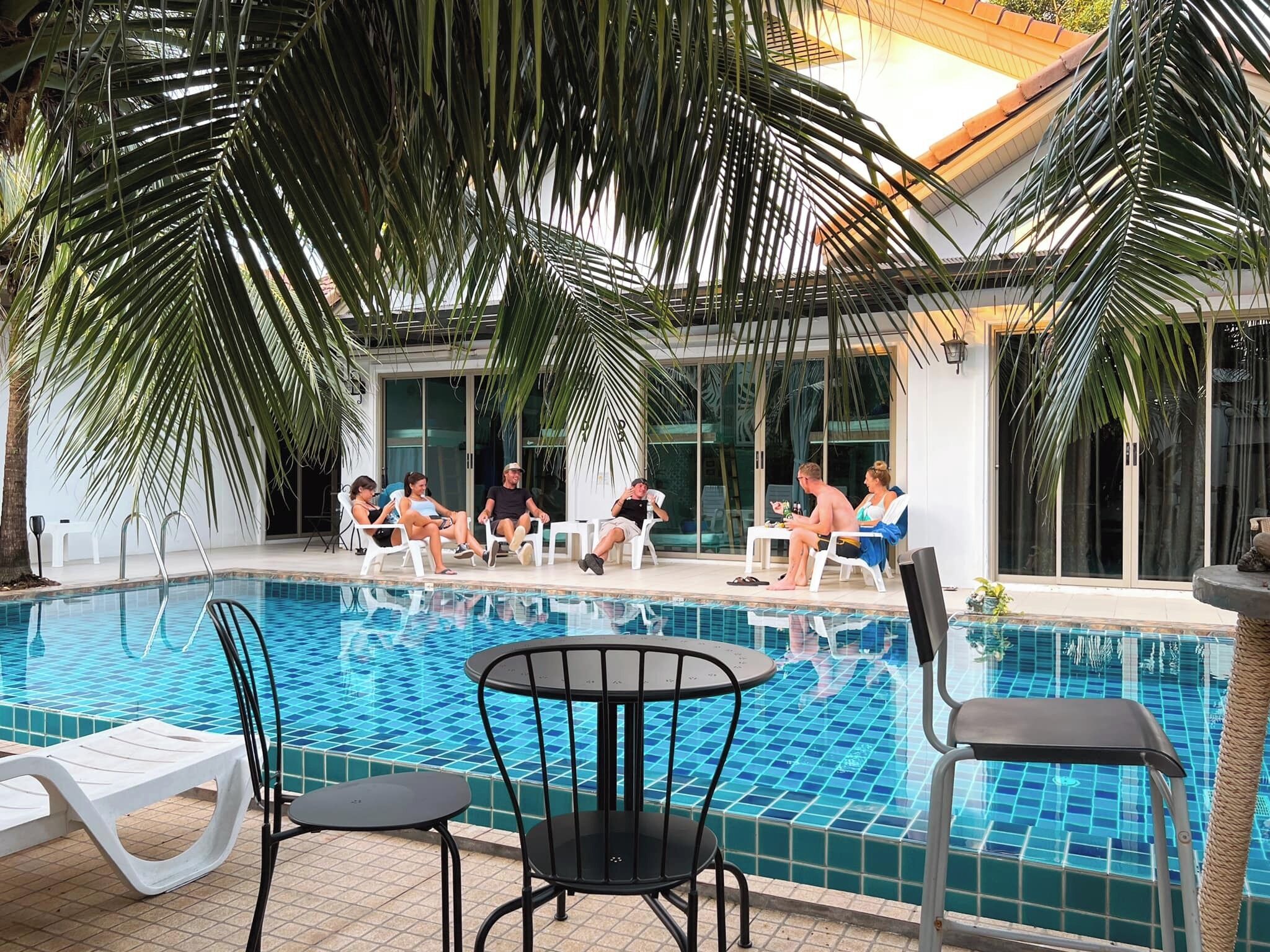 Uma área à beira da piscina com azulejos, espreguiçadeiras e palmeiras.