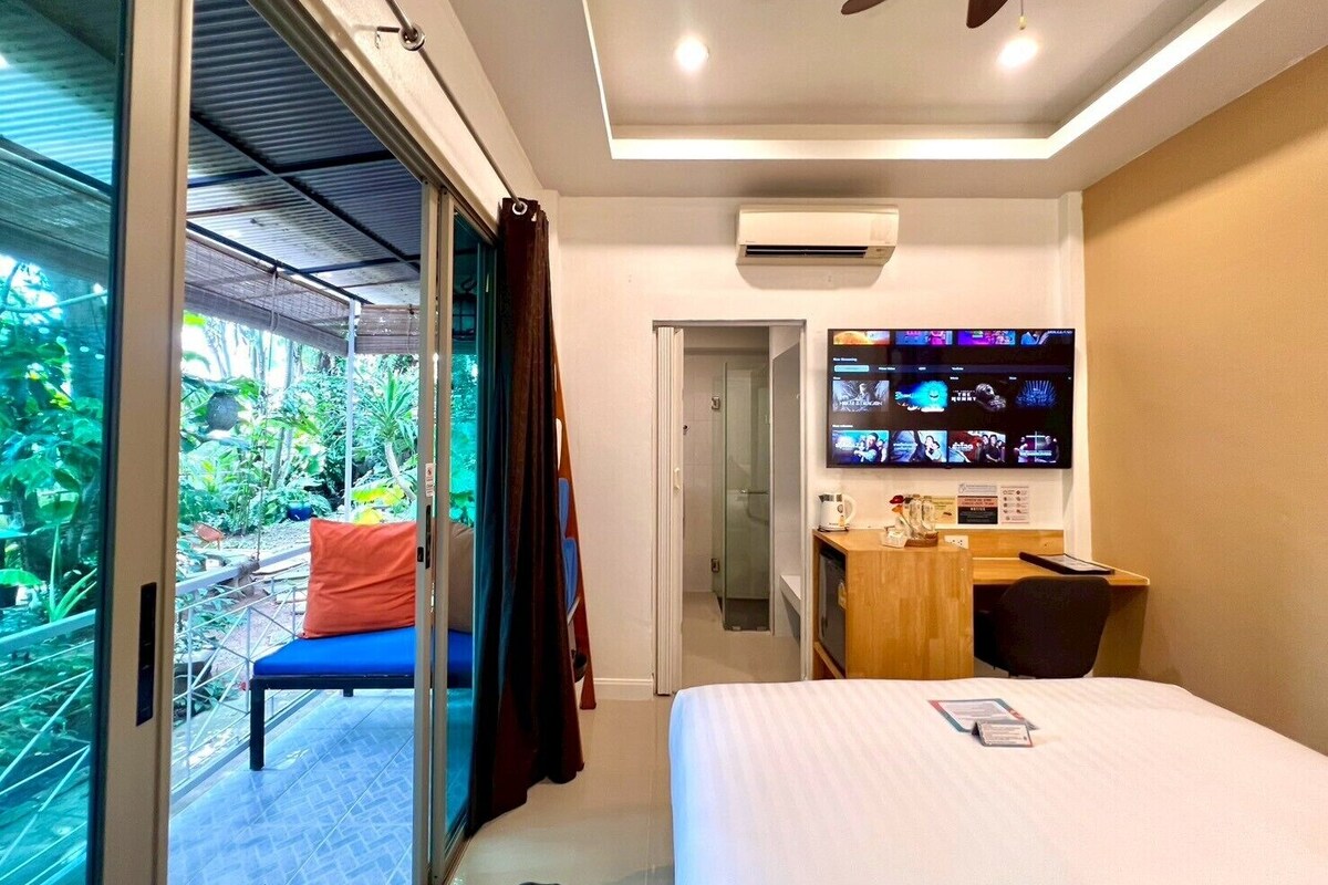 A modern hotel room with a bed, a desk, a TV, and a sliding glass door leading to a balcony with plants.