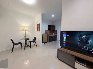 A modern living room with a flat-screen TV, a dining area with chairs, a lamp, and a kitchen in the background. - The Snug Airportel (Sa Khu)