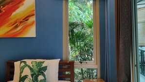 A bedroom with a bed, a nightstand, a window with a view of greenery, and a ceiling light fixture. - The Snug Airportel (Sa Khu)