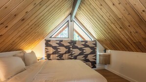A cozy attic bedroom with a sloped wooden ceiling, a large bed, a window with a view, and a patterned wall.