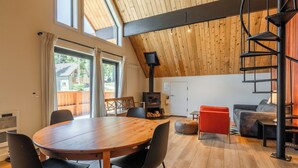 A modern living room with a wooden ceiling, a round wooden table with chairs, a sofa, and a red ottoman.
