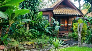 Une maison rustique en bois, avec un toit de tuiles, entourée d’une végétation tropicale luxuriante et traversée par un petit ruisseau.