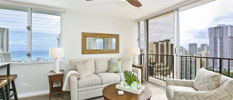 A spacious living room with a large window, a ceiling fan, a sofa, a patterned armchair, and a coffee table.