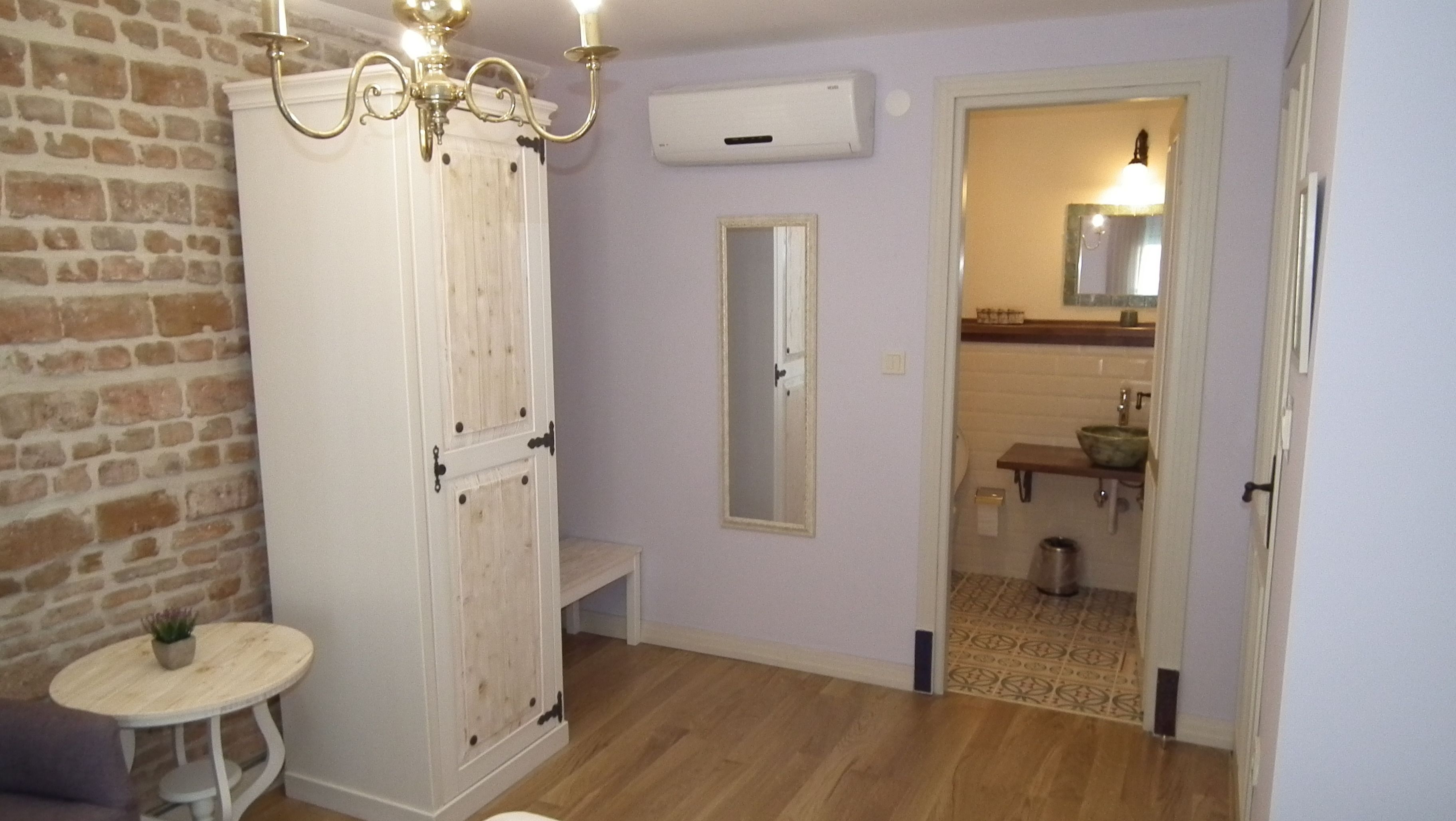 Ein Zimmer mit Backsteinwand, einem weißen Kleiderschrank, einem kleinen runden Tisch mit Pflanze, einem Spiegel und einem durchgehenden Türbogen sichtbarem Badezimmer.