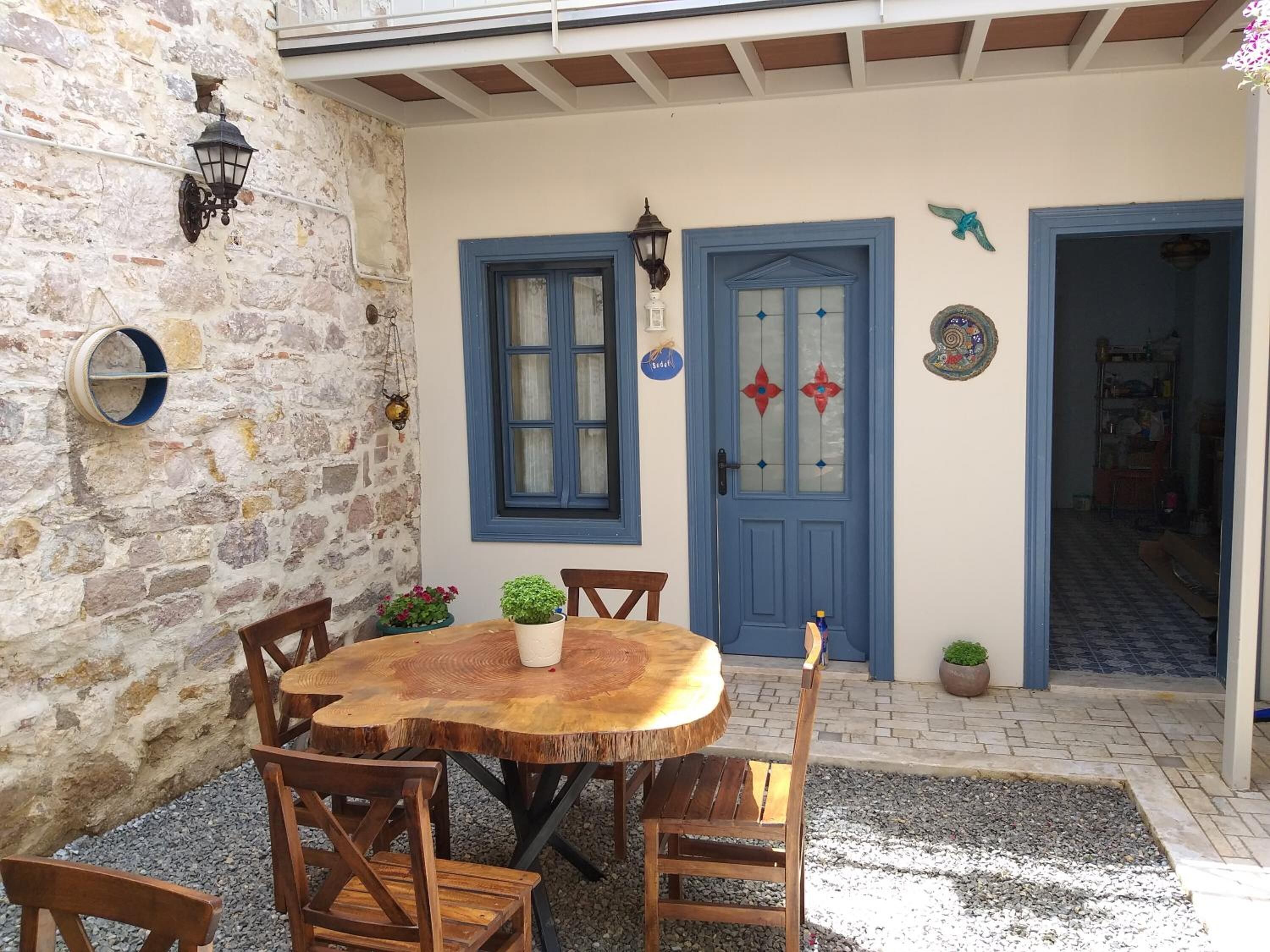 Un muro de piedra, una mesa redonda de madera con sillas, una puerta azul con paneles de vidrio y un pequeño jardín.