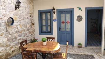 Un muro de piedra, una mesa redonda de madera con sillas, una puerta azul con paneles de vidrio y un pequeño jardín.