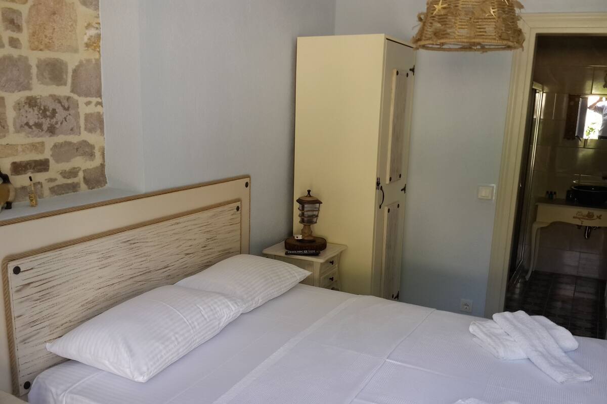 A neatly made bed with white linens and towels, a wooden headboard, a bedside lamp, and a stone wall.