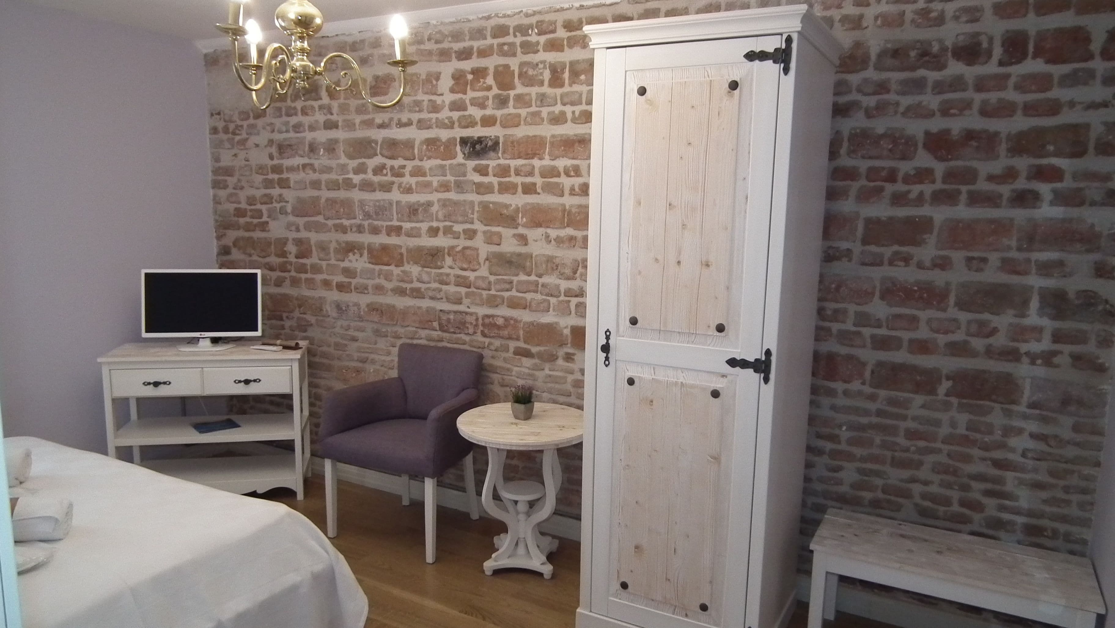Een kamer met een bakstenen muur, een witte garderobe, een ronde tafel met een plant, een paarse stoel, een witte nachtkastje met een televisie en een bed met witte lakens.