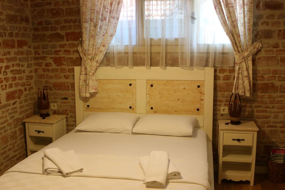 A room with a wooden headboard bed, white linens, a small bedside table with a lamp, and a brick wall.