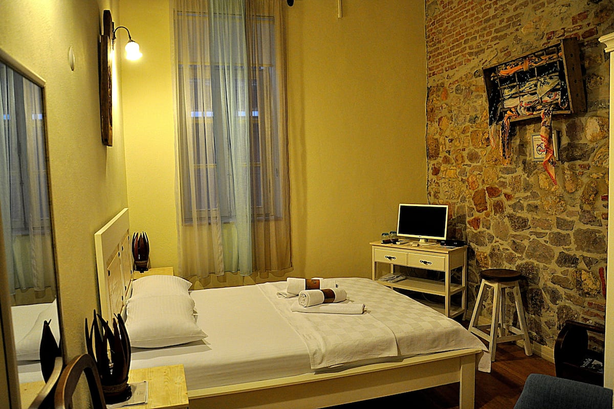 A hotel room with a bed, a desk with a computer, a mirror, and a stone wall.