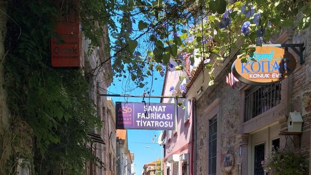 A narrow cobblestone street lined with buildings, some with signs indicating local businesses. - Gizemli Konak (Ayvalik)