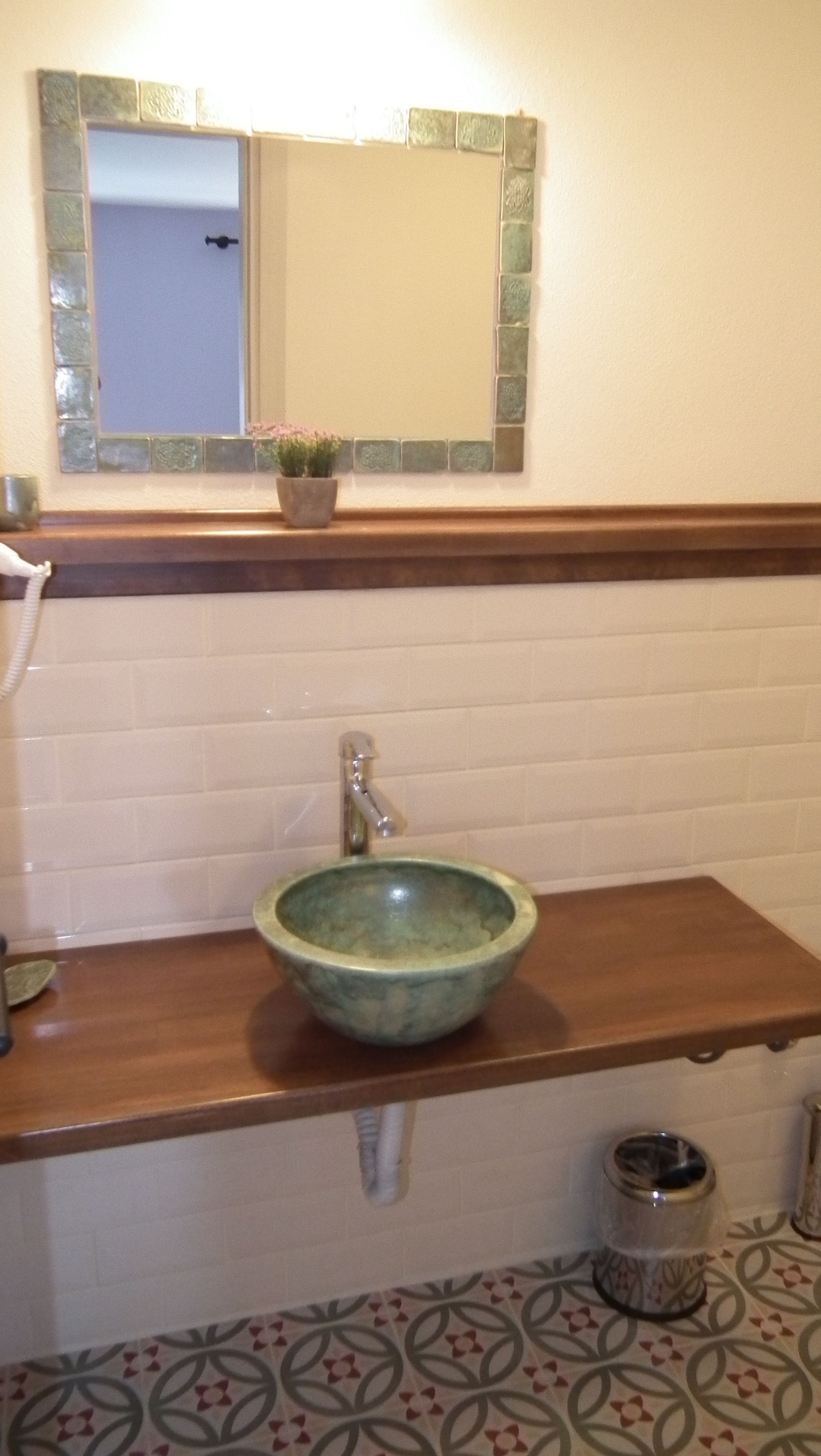 Un baño con tocador de madera, lavamanos de cerámica verde, pared con azulejos blancos y un espejo con marco verde.